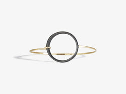 A blackened sculptural circle is attached to a forged 14k yellow gold bangle that hooks into the circle